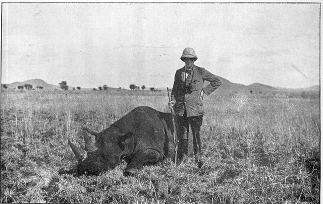 A hunter stand over the body of a white rhino in an old, black & white photograph