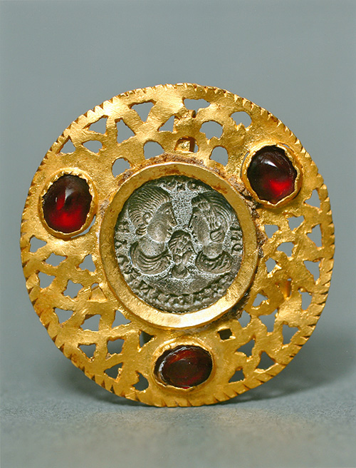 A gold pendant flanked with red gems with a silver inset depicting three human figures.
